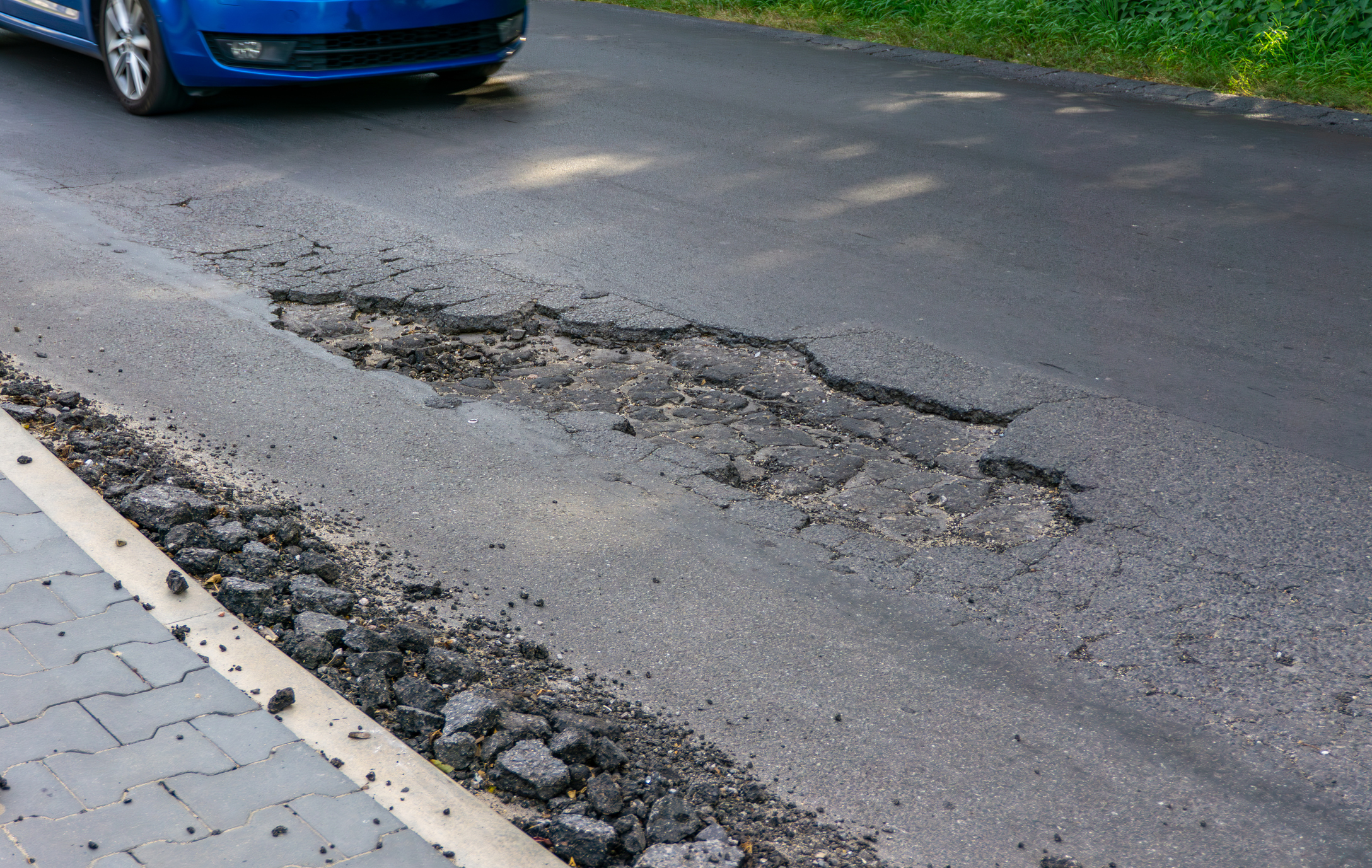 Car driving towards a large pothole in asphalt near curb, visible surface damage and broken road edges.